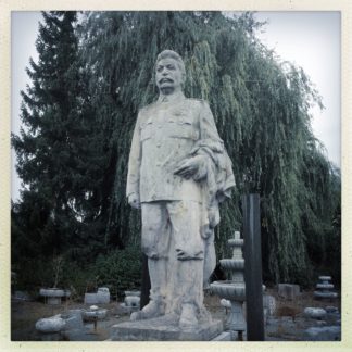 Lebensgroße Stalin-Skulptur auf Sockel vor Nadel- und Laubbaum. Schwarz-weiß-Bild.