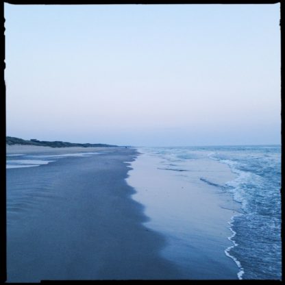 Auf der Linie der ankommenden seichten Wellen stehend blickt man am Strand entlang. Grau-blauer Farbton und blasser HImmel