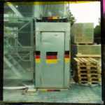Eine Blechtür mit aufgemalten Deutschland-Farben in einem Bauzaun. Daneben ein Stapel Euro-Paletten.
