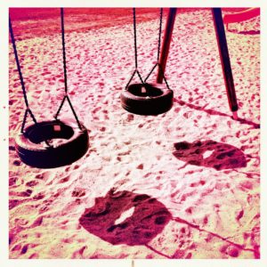 Zwei alte Autoreifen, umfunktioniert zur Kinderschaukel werfen Schatten auf Sand. Rötlich verfärbtes Bild.