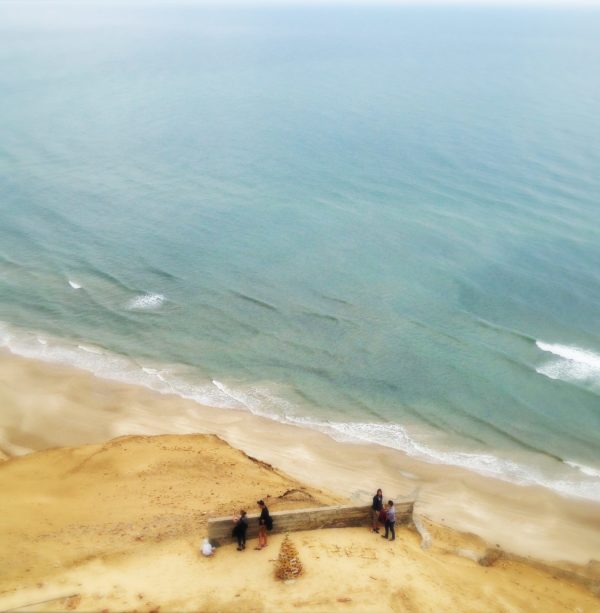Schräg von links nach rechts zieht sich die Küstenlinie der ruhigen Nordsee vor einem steilen Sanddünenhang. Aus der Vogelperspektive blickt man auf winzige Menschen, die auf einer zerfallenden Mauer ruhen.