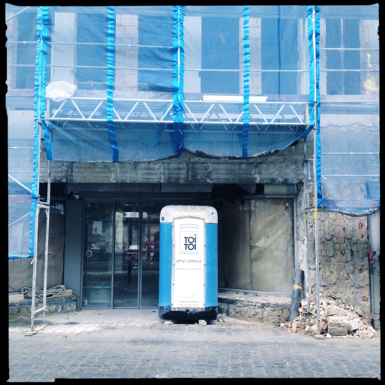 Ein Toi Toi Mietklo vor einer Baustelle. Das Portal wirkt wie eine Bühne. Rechts und links ist das Baugerüst mit Netzen abgehängt.