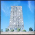 ein silbrig schimmerndes Hochhaus mit stürzenden Linien ragt in den blauen Himmel. Am unteren Bildrand ein paar Bäume.
