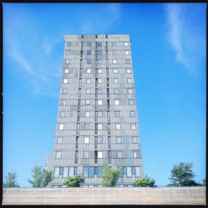 ein silbrig schimmerndes Hochhaus mit stürzenden Linien ragt in den blauen Himmel. Am unteren Bildrand ein paar Bäume.