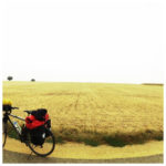 Vor einem abgeernteten Getreidefeld steht ein bepacktes Reiserad. Die Gegend ist flach bis zum Horizont.