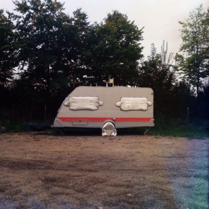 Ein Wohnwagen im Profil vor einer dunklen Hecke. Fenster und Rad sind verhängt, was ihm ein Gesicht gibt.
