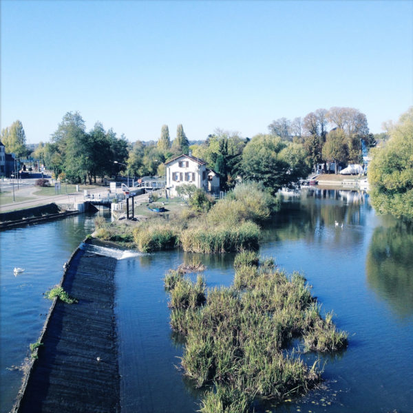 Bläuliches Bild von einer Brücke hinab auf einen Fluss. Ein Schleusenhaus und ein Stauwehr schmiegen sich an Uferbewuchs.