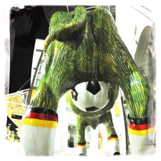 Blick underfoot auf den Hintern einer kunststoffenen Kuh. Das Euter ist ein Fußball. Sie trägt Socken mit Deutschlandfarebnbund. Ihr Körper ist grün.
