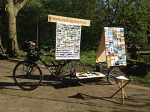 Fahrrad mit Anhänger, auf dem ein fast zwei Meter hohes Display mit vielen kleinen quadratischen Bildern steht. Ein Schild mit der Aufschrift www.radelgalerie.de dient als Aufgängung für ein Plakat über dem Fahrradsattel