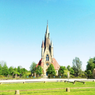 Reichlich verzierter Kirchturm ragt aus flachem Land in Cyanblauen Himmel. Das Grün junger Linden garniert die Friedhofsmauer.