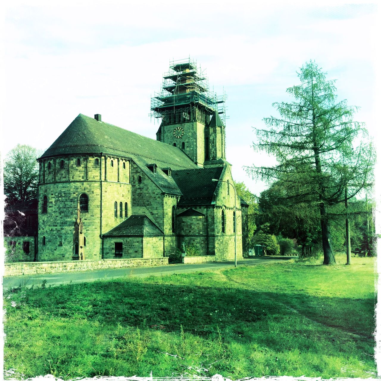 von der Rückseite blickt man auf ein Kirchenschiff. Das Bild ist grünlich verfremdet. Der Kirchturm ist von einem Gerüst umgeben. Neben der Kirche rechts steht ein Nadelbaum, der fast so hoch ist wie der Turm.