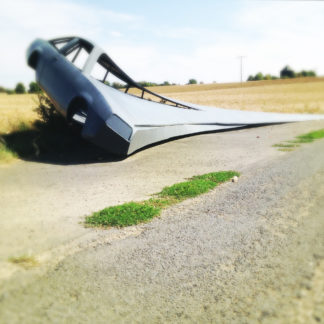 Seitenteil eines Opel Manta, das mit einer stählernen Schwinge verschweißt auf einem abgeernteten Feld steht. Die Schwinge sieht aus wie der Flügel eines Teufelsrochens.