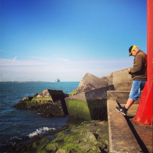 An einem roten Mast rechts im Bild lehnt ein Fotogrf mit Schirmmütze. Hinter Betonblöcken öffnet sich das Meer. Schiffe und Hafenanlagen am Horizont.