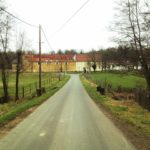 Blick eine schmale Straße entlang , die auf einen französischen Hof zuläuft. Die aneinander gebauten Gebäude zeigen eine klare Linie der Trennung durch unterschiedliche Farben, gelb und weiß. Ringsum Wald und Weiden.