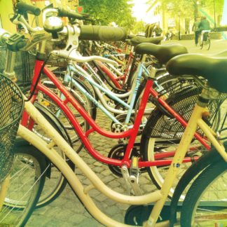 Nahaufnahme durch die Rahmen vieler Fahrräder, die aufgereiht an einem Fahrradparkplatz setehen. In dem gelblich verfärbten Bild sieht man rechts im Hintergrund Menschen und Radler.