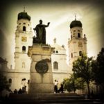 Schwarz-weiß Retro Bild des Passauer Doms, an dessen Front ein dunkles Denkmal steht. Eine Vignettierung rahmt das Bild. Zwei Kuppelkirchtürme, Bäume und Touristen.