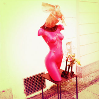 Eine lila Schaufensterbüste ohne Kopf und ohne Beine. Farbverfremdetvor gelblicher Hauswand und sparsam geschmückt.