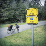 zwei etwa Din A 4 große gelbe Radwegschilder an einem Pfosten vor dem bepackten Fahrrad im Wald