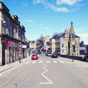 Straßenszene in der schottischen Stadt Tain. Mittig fluchtend blickt man eine Straße entlang. Ein rotes Auto schiebt sich ins Bild, gothisch anmutende Zierart an den bleichen Hauswänden.