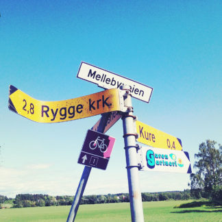 Schildergewirre in blassblau mit gelb. Ein verbogenes Schild zeigt nach Rygge KRK, der Rygge-Kirche in Norwegen.