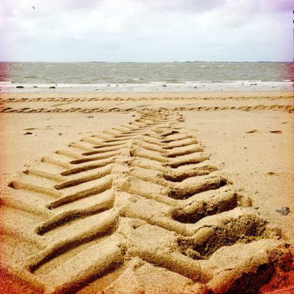 Eine Traktorspur führt gezackt durch Sand aufs Meer zu