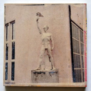 eine heroische Figur in Pastellfarben fast weiß vor der Fassade eines Wasserkraftwerks.