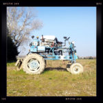 Eine blaue Maschine, die einem hochgelegten Traktor ähnelt auf weitem Feld. Links im Bild ein Gebüsch.
