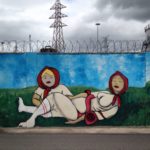 Vor einem etwa ein drittel des Bilds einnehmenden Himmel mit dunklen Wolken und Strommast ist auf einer Stacheldraht bewehrten Mauer ein Graffiti mit zwei liegenden, schlummernden Matruska-ähnlichen Frauen gesprayt.