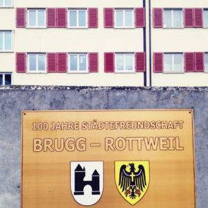 Vor einem Wohnhaus mit roten Fensterläden hängt an einer Betonwand ein bräunlich-goldenes Schild mit der Aufschrift Stätdepartnerschaft Brugg Rottweil. Darunter die Wappen der beiden Städte.