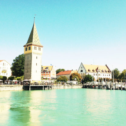 Cyanfarben bis weiß ist die Szene des Hafens von Lindau, betrachtet vom Boot aus über Wasser. Markant ragt ein Turm im linken Teil des Bilds.