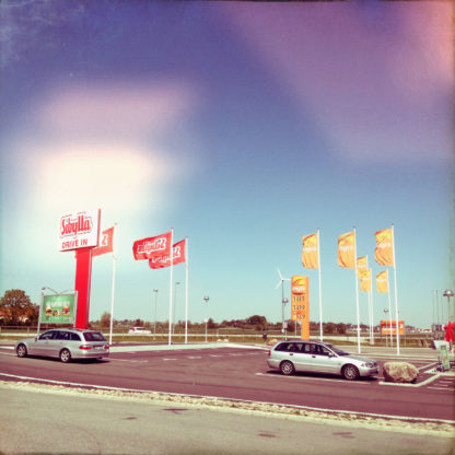 Ein Bild mit gelb-rosa Fehlfarbeneinflüssen zeigt einen Autorastplatz mit PKW, Tankstellenschild und Fahnen.