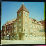 Ein stattliches Eckhaus mit quadratischem Türmchen als verfärbtes Bild im Retrostil. Der Bildrand ist schwarz-rosa-gelblich.