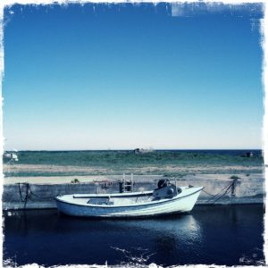 Längsseits betrachtet ein kleines, weißes Boot ohne Mast, das an einer Hafenmauer liegt. Der blaue Teint des Bildes zieht sich vom Hafenbecken über die Wiese bis ins offene Meer und den Himmel.