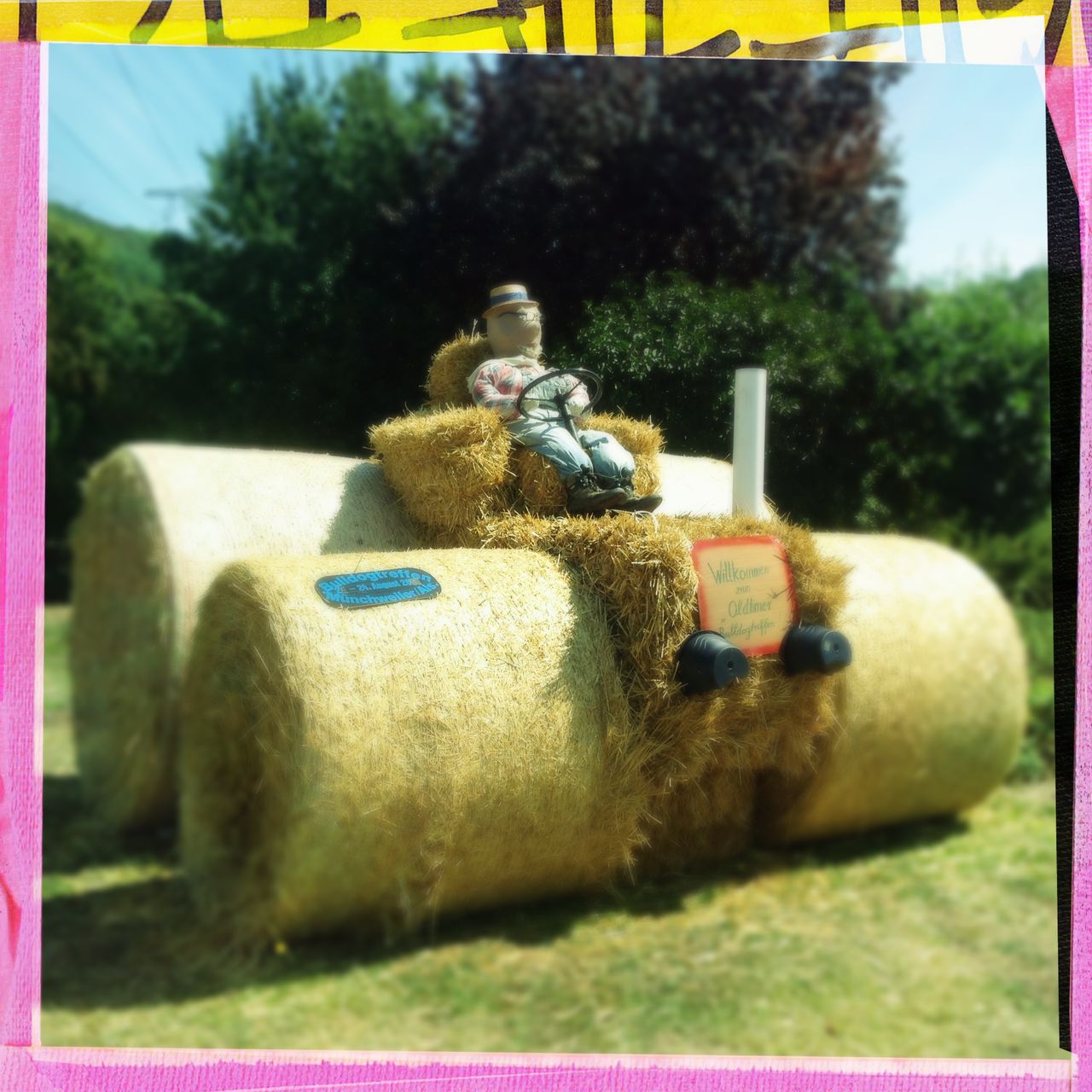 Ein Traktor aus Stroballen mit riesigen Rädern und einer Puppe als Traktorist. Das Bild hat einen gelb-rosa Rahmen.