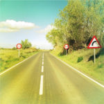 Ein im Siebziger-Jahre typischen Fotostil mit gelblich-türkisen Fehlfarben fotografiertes Retrofoto einer spanischen Landstraße. Zwei Warnschilder rechts und eines links zeigen eine Rechtskurve an.