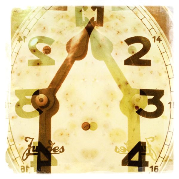 Eine gespiegelte Uhr im Detail mit altem Zeiger. Das Überblendete Bild zeigt die Ziffern 1 bis 4. Der kleine Zeiger steht auf 1, der große endet ber dem Schriftzug Juges (verdeckt ihn zum Teil. Gelblich beige Farbe.