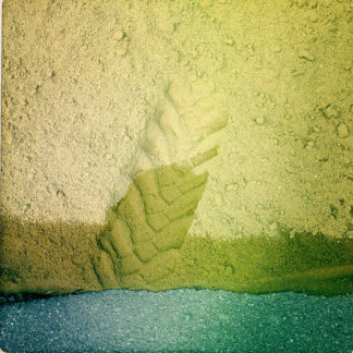 Von oben blickt man auf einen Haufen Sand, der auf Teer zu liegen kommt, klare Horizontlinie zwischen Teer und dem gelb des Sandes im unteren Bilddrittel. Mittig führt die Spur eines Traktorhinterreifens wie ein französischer Akzent in den Sand. Dort wo der Sand den Teer berührt ist er nass und somit etwas dunkler.