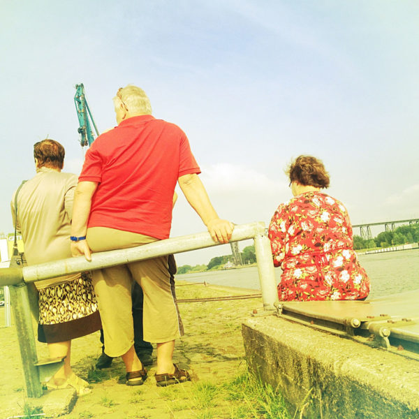 In dem retro gelblich rötlich verbleichten Bild sitzen und stehen drei Menschen mit dem Rücken zum Betrachter und beäugen den Hafen am Nordostseekanal