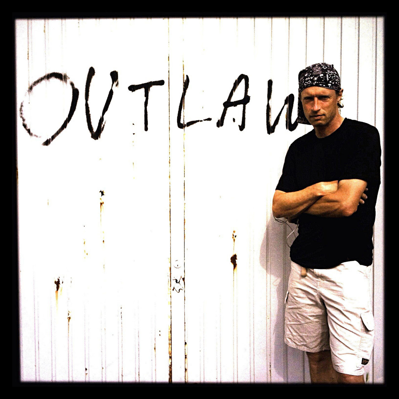 Mann mit schwarzem T-Shirt und kurzer weißer Hose, verschränkten Armes vor einer weißen Wand, an der das Wort Outlaw gesprayt ist