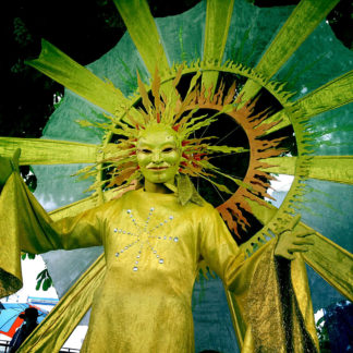 Ein als Sonne verkleideter Mensch mit goldgelbem Kostüm und ausschweifenden Strahlen schaut von oben in die Kamera.