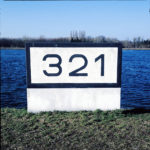 Ein schlichter Betonklotz, weiß, mit schwarzem Rand vorm tiefblauen Fluss. Die Rheinkilometrierung 321 steht in serifenlosen Buchstaben darauf.