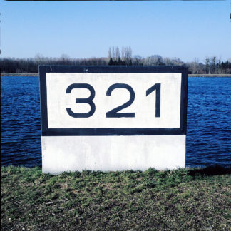 Ein schlichter Betonklotz, weiß, mit schwarzem Rand vorm tiefblauen Fluss. Die Rheinkilometrierung 321 steht in serifenlosen Buchstaben darauf.