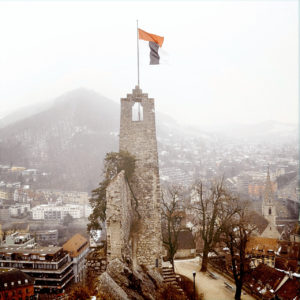 Ein schmaler Burgturm über einer kleinen, vernebelten Stadt. Am Bergfried weht eine Fahne.