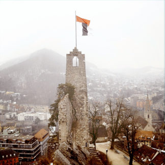 Ein schmaler Burgturm über einer kleinen, vernebelten Stadt. Am Bergfried weht eine Fahne.