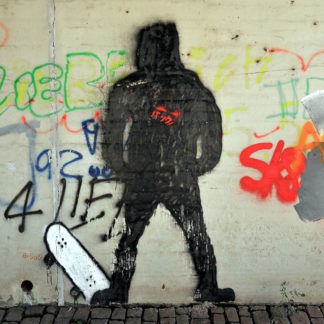Eine Graffiti-Wand mit Hauptmotiv der schwarzen, gesprayten Silhouette eines vermummt wirkenden Skaters, der den Fuß links auf einem aufgestellten Skateboard zeigt. Daneben zahlreiche Tags und Schriftzüge.
