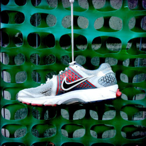 Ein Turnschuh der Marke Nike an einem grünen Kunststoff-Waben-Bauzaun hängend.