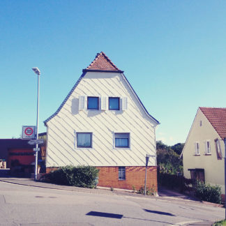 Ein Haus mit Krüppelwalmdach und vier großen und einem kleinen Fenster steht an einer von links nach rechts fallenden Straße.