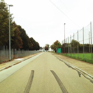 In der Seitenstraße zwischen dem hohen Zaun eines Fußballplatzes und einem niedirgen Zaun links gegenüber endet eine pechschwarze Bremsspur im unteren Bilddrittel.