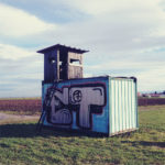Auf einem mit Graffiti bemalten Container thront ein hölzerner Jagshochsitz.
