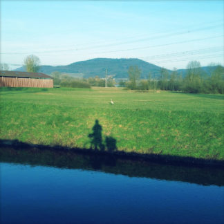 Über einen Kanal hinweg schaut man in dem bläulich-grünen Bild auf den Schatten eines Radlers an der Uferböschung. Im Hintergrund mittelgebirgsartige Berge.